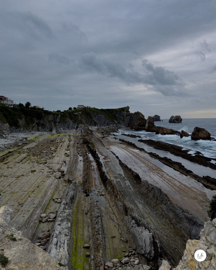 Estratos rocosos inclinados en la costa de Arnía bajo cielo nublado.