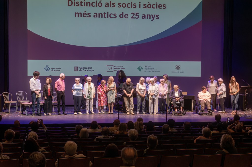 Escenario del Teatro Monumental de Mataró durante el acto del 45 aniversario de Aules Sènior, con los socios homenajeados sobre el escenario y el público observando desde las butacas.